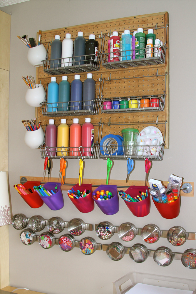 A corkboard is on a wall with shelves with paints and other art supplies. A row of small magnetic containers is also attached to the wall and filled with small art supplies like paper clips, etc. 